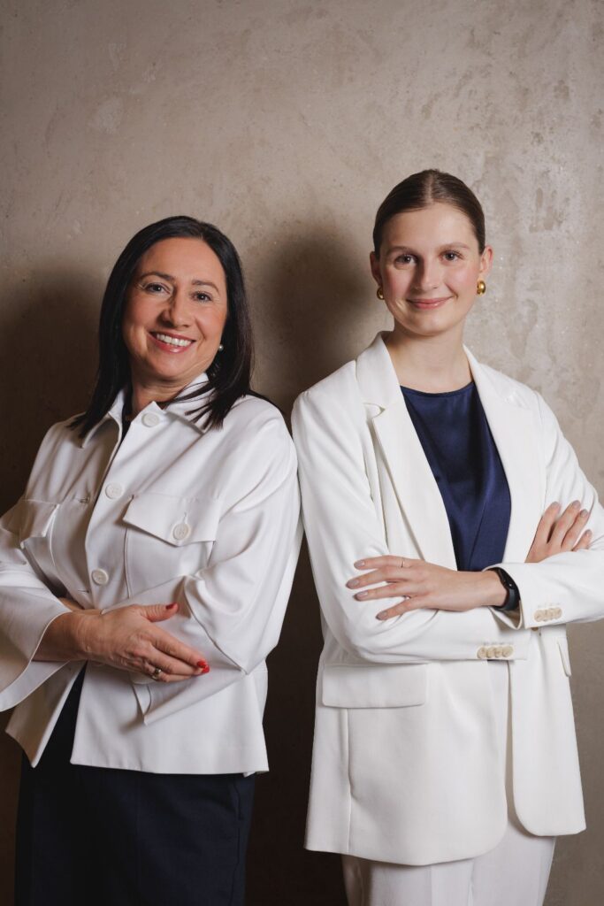 Zwei Geschäftsfrauen in eleganten weißen Blazern stehen nebeneinander und lächeln in die Kamera, eine symbolisiert reife Erfahrung, die andere jugendliche Dynamik.
